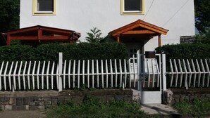 A two-story building with a white exterior, two small windows with yellow frames, and a wooden porch on the ground floor.