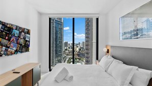A modern hotel room with a large bed, a wooden console table, a wall-mounted TV displaying movie posters, and a city view through floor-to-ceiling windows. - Luxury Condos in Downtown Miami (Miami)