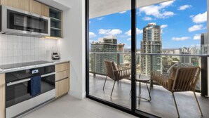 A modern kitchen with a built-in microwave and oven, a balcony with two chairs and a table, and a city view through large windows. - Luxury Condos in Downtown Miami (Miami)