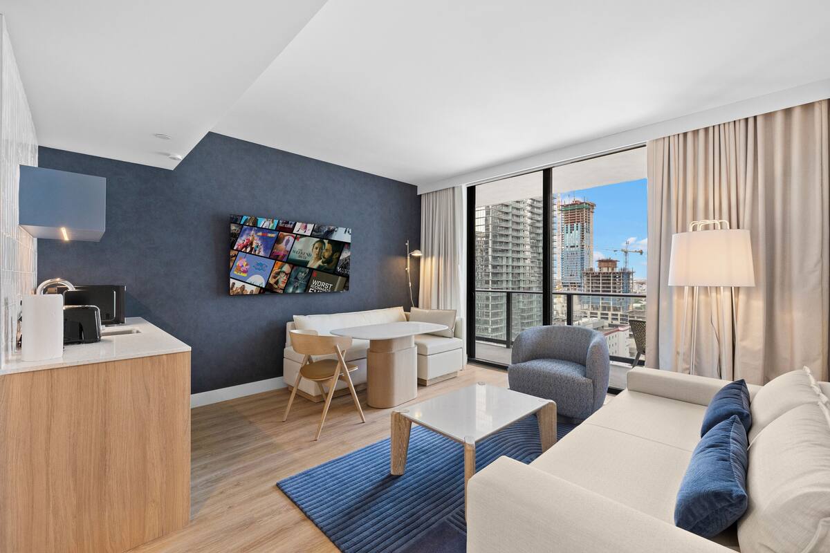 A modern hotel room with a large window, a dining area, a sofa, and a TV mounted on the wall.