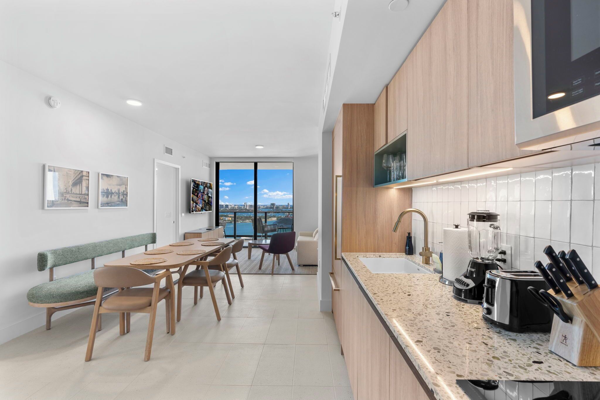A modern kitchen with a granite countertop, a built-in oven, and a sink. There is a dining area with a table and chairs, and a bench along the wall.