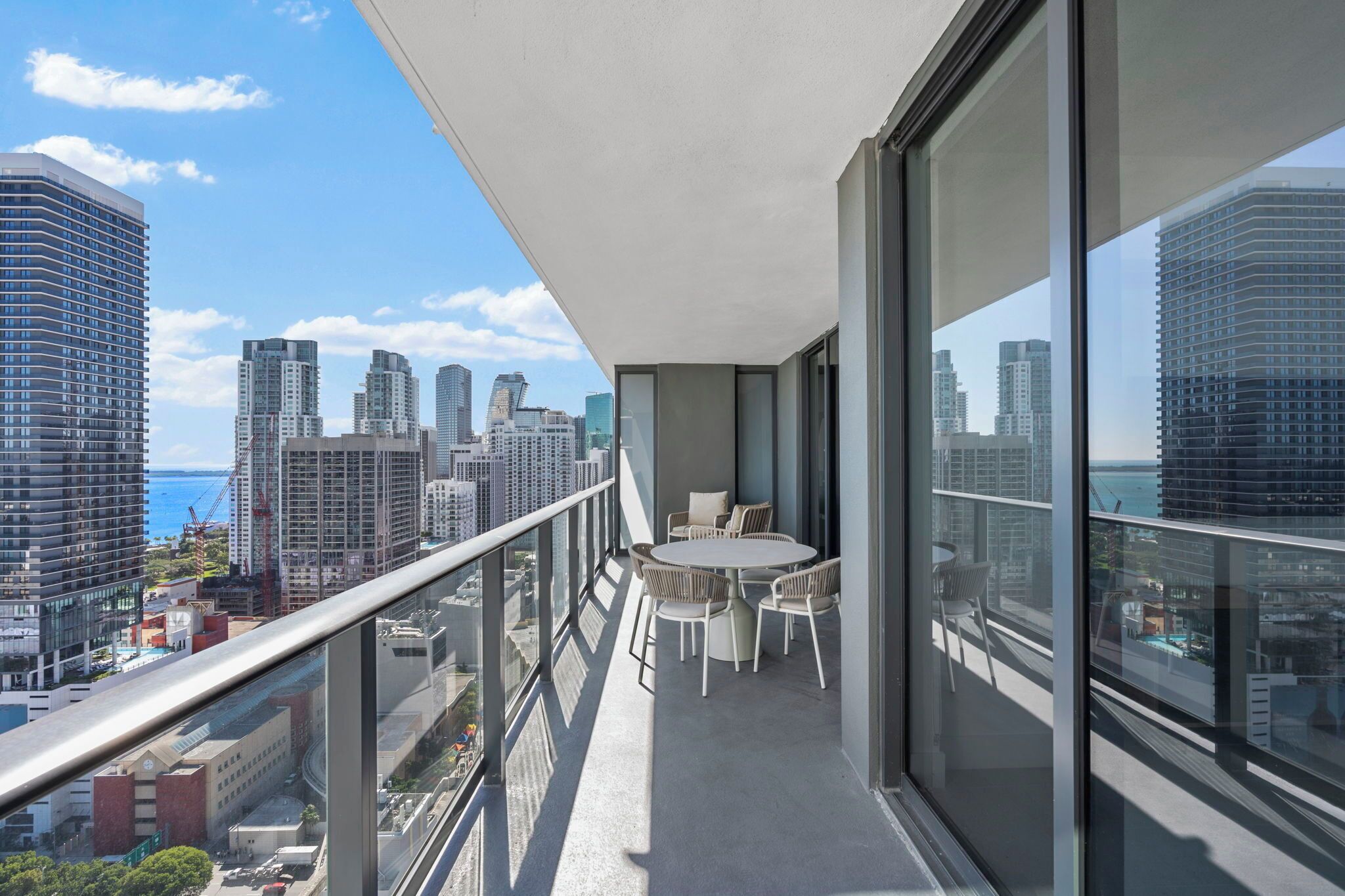 A balcony with a table and chairs, overlooking a cityscape with tall buildings and a view of the ocean.