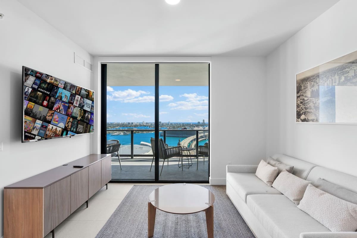 A modern living room with a large TV, a sofa, a coffee table, and a balcony with a view of the sea.