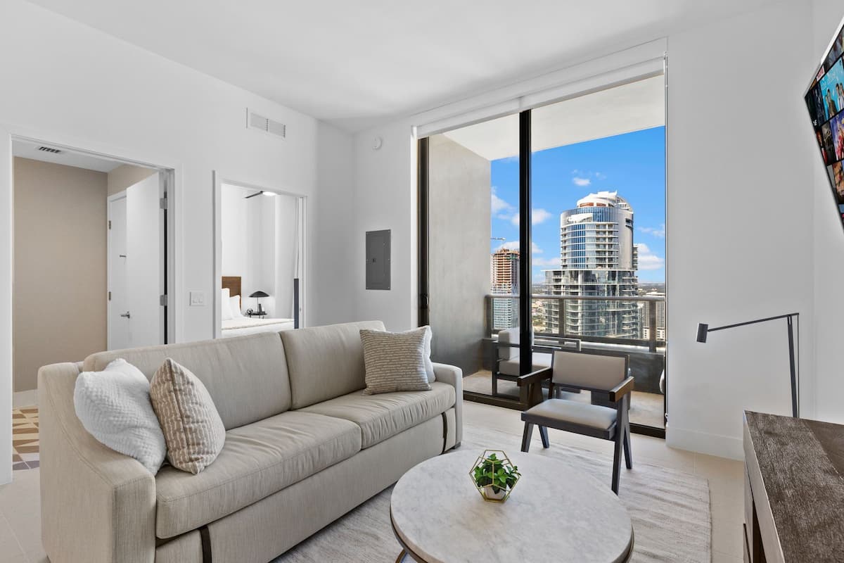 A modern living room with a sofa, a coffee table, and a view of a cityscape through large windows.