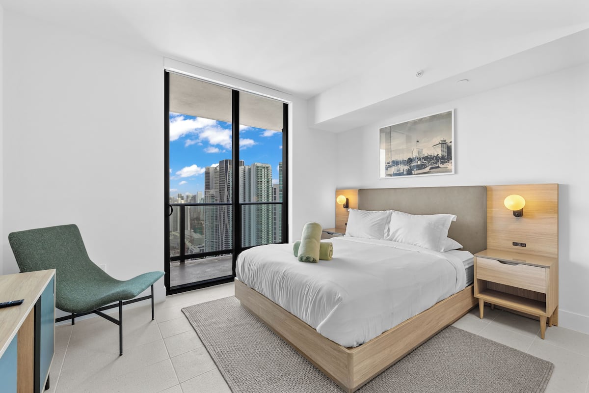 A modern hotel room with a large bed, a desk, a chair, a nightstand, and a view of the city skyline through a sliding door.