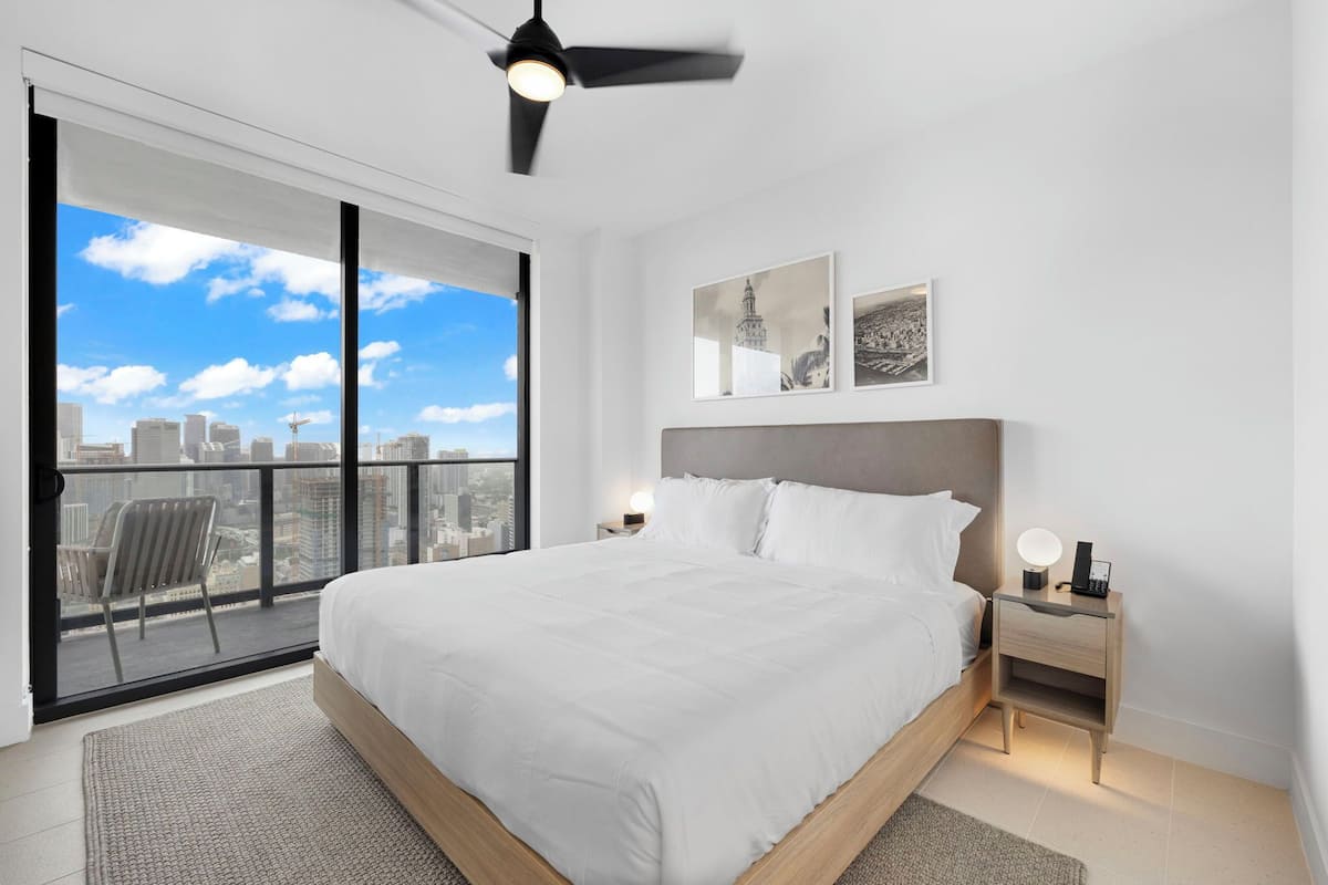A modern bedroom with a large bed, a city view balcony, and a ceiling fan.