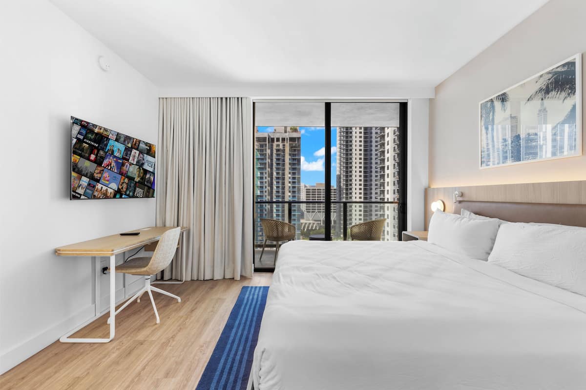 A modern hotel room with a large bed, a desk, and a view of high-rise buildings.