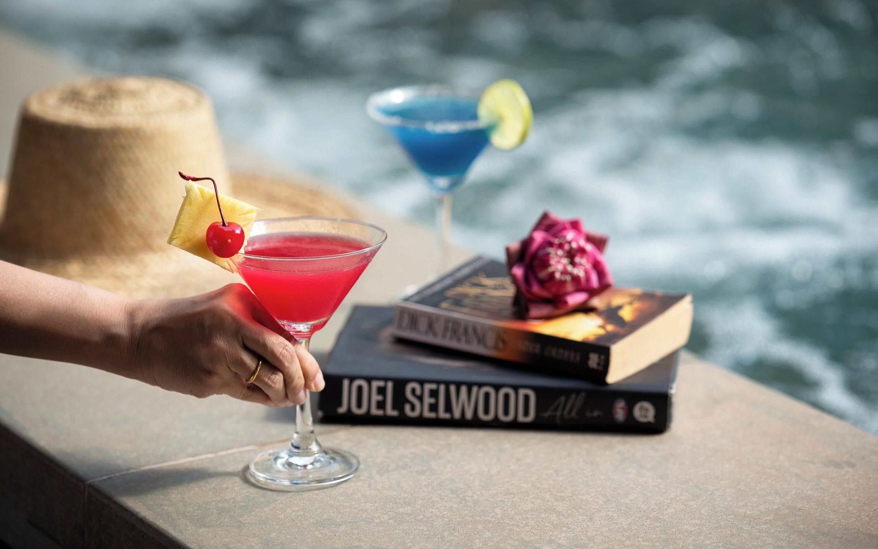 Una mano sostiene una copa de martini con una cereza y una rodaja de lima, un cóctel azul con adorno de lima, y un libro titulado 'JOEL SELWOOD' sobre un saliente junto al mar.