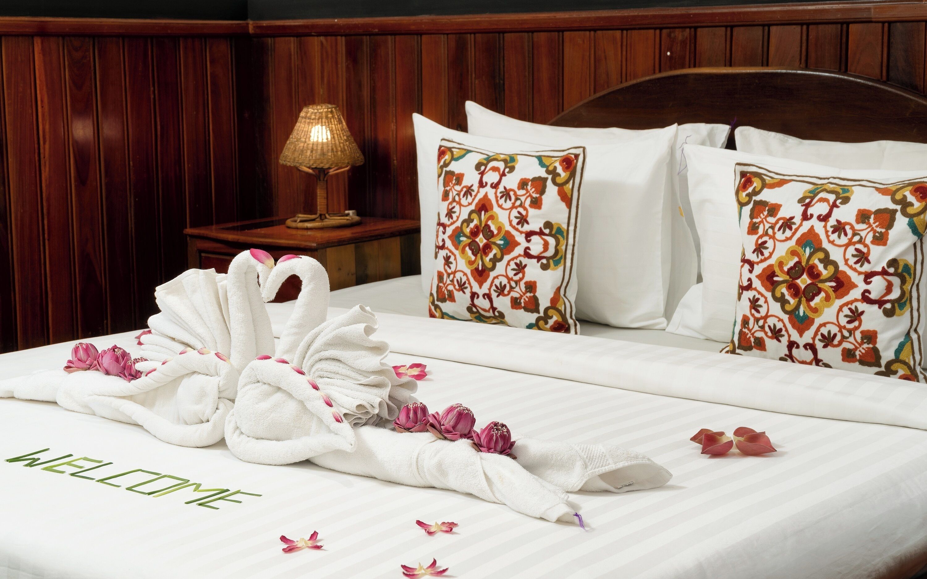 Een netjes opgemaakt bed met een handdoek gevouwen in de vorm van een zwaan, kussens met bloemenmotieven en een bord met de tekst 'WELCOME' op het bed.