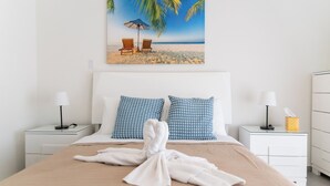 A neatly made bed with a towel, two blue patterned pillows, and a beach scene painting above it.