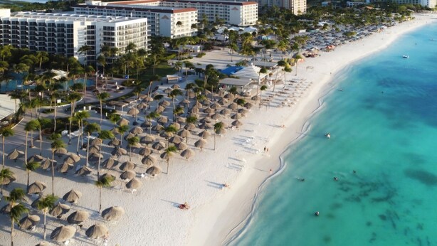 An aerial view of a beach resort with multiple hotel buildings, palm trees, and beach umbrellas.