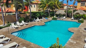 A swimming pool with white lounge chairs, palm trees, and a thatched umbrella.