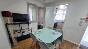 Un espacio habitable pequeño con una mesa de comedor, sillas, un televisor sobre un soporte y una cocina visible a través de una puerta abierta.