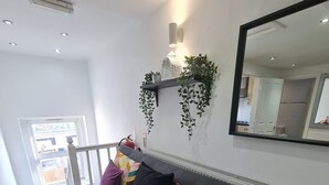 Una sala moderna con un sofá gris, cojines coloridos, un estante montado en la pared con plantas, un espejo grande y vistas a la cocina a través de una puerta abierta.