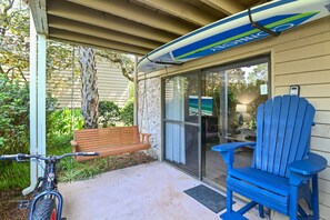 Une terrasse couverte avec un banc, une chaise Adirondack bleue, une planche de surf et un vélo.