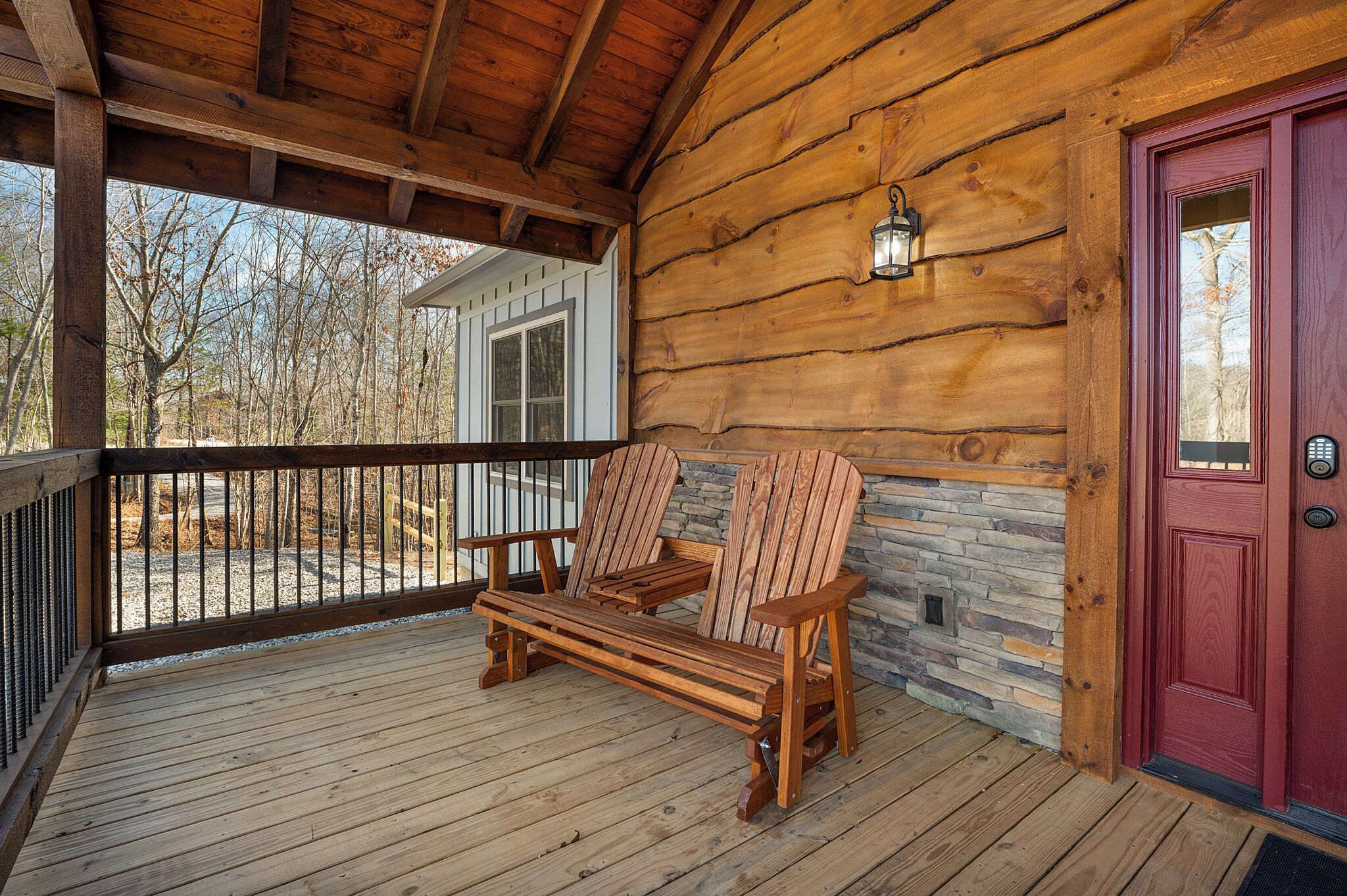 A wooden porch with a bench and chairs, a stone wall, and a red door.