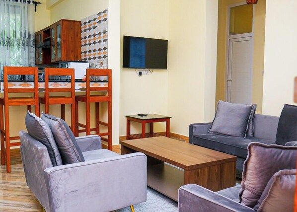 A living room with a sofa, armchairs, a coffee table, a TV, and a dining area with chairs and a table. - VvEK BED AND BREAKFAST APARTMENT (Arusha)