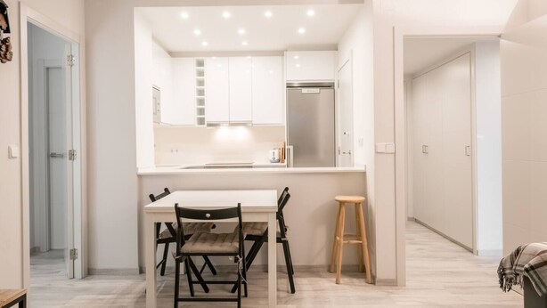 Cozinha moderna com armários brancos, geladeira de aço inoxidável e área de jantar com mesa e cadeiras.
