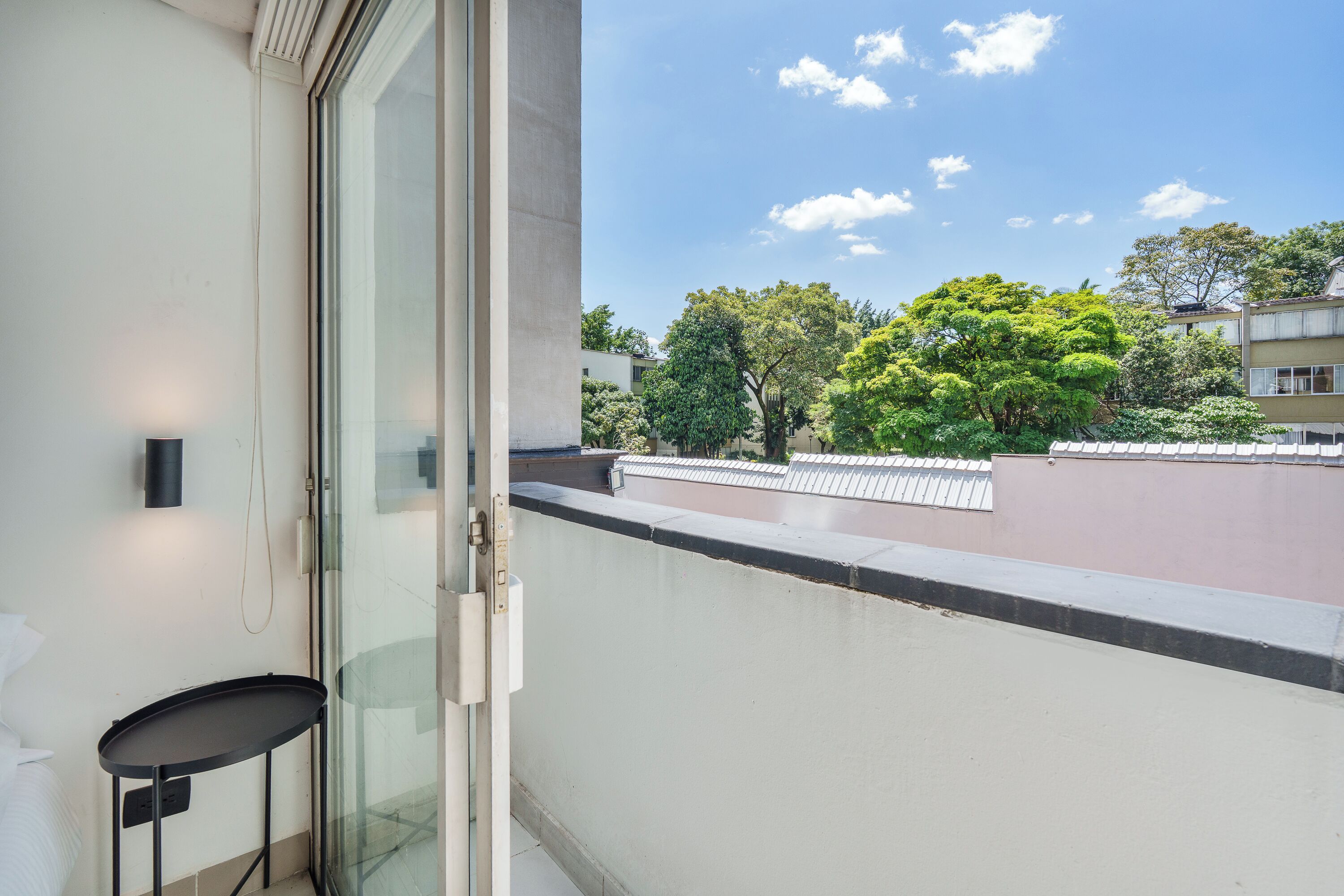 Un balcón con puerta de vidrio, una mesa pequeña y vistas a árboles y edificios.