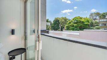 Un balcón con puerta de vidrio, una mesa pequeña y vistas a árboles y edificios.