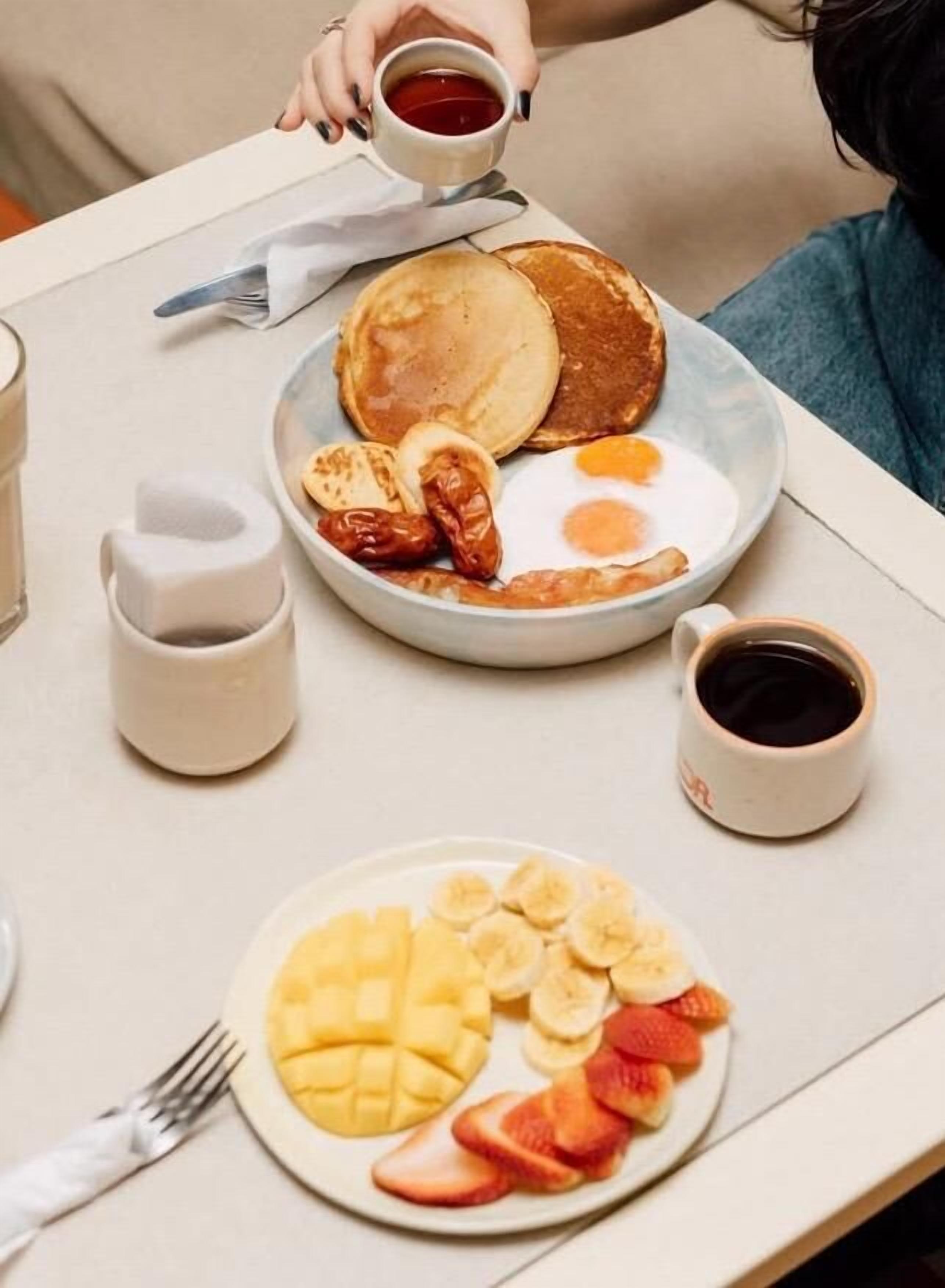A breakfast setting with pancakes, eggs, and fruit on plates, along with cups of coffee and tea.