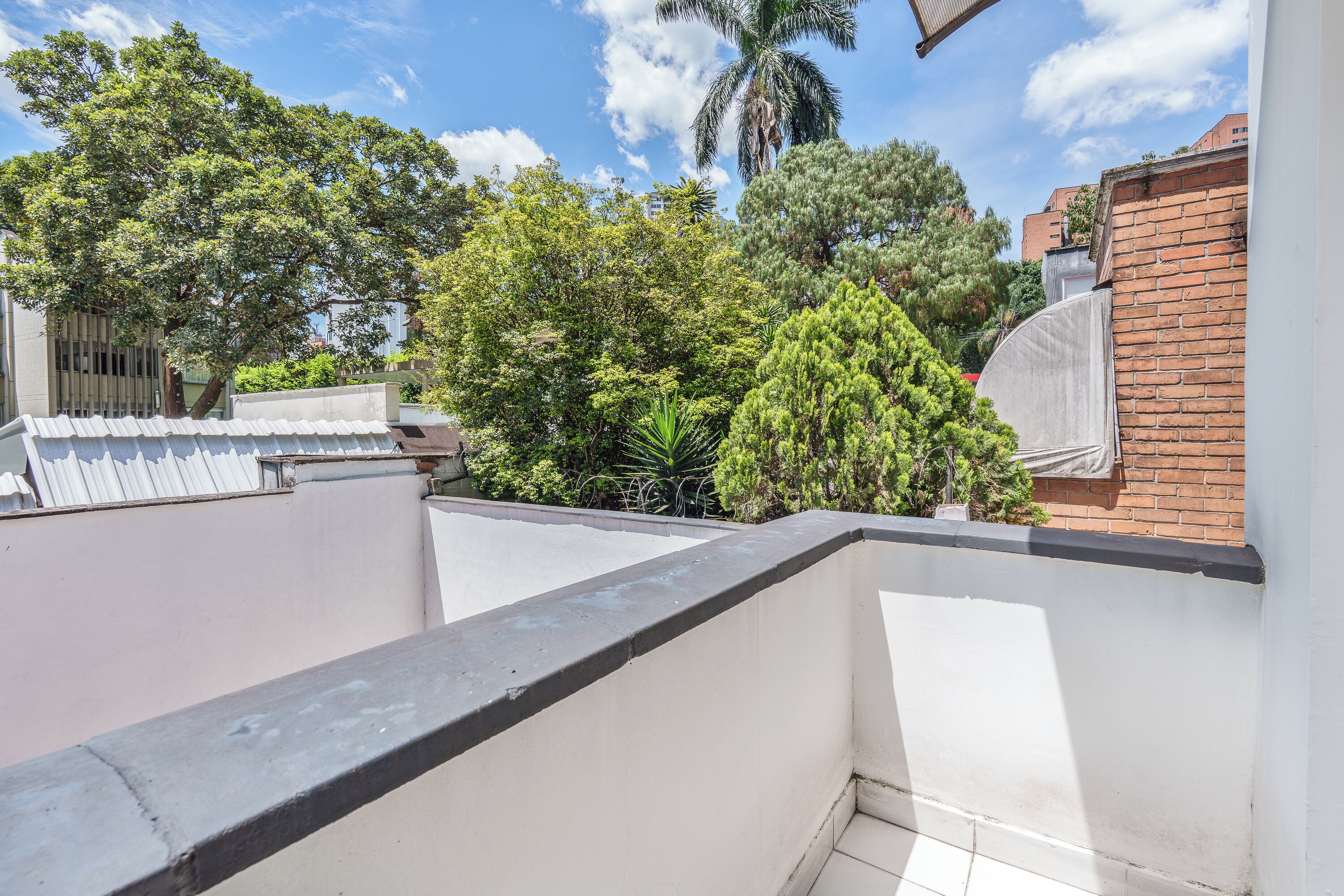 A rooftop terrace with a white railing, a view of trees, and a brick wall.