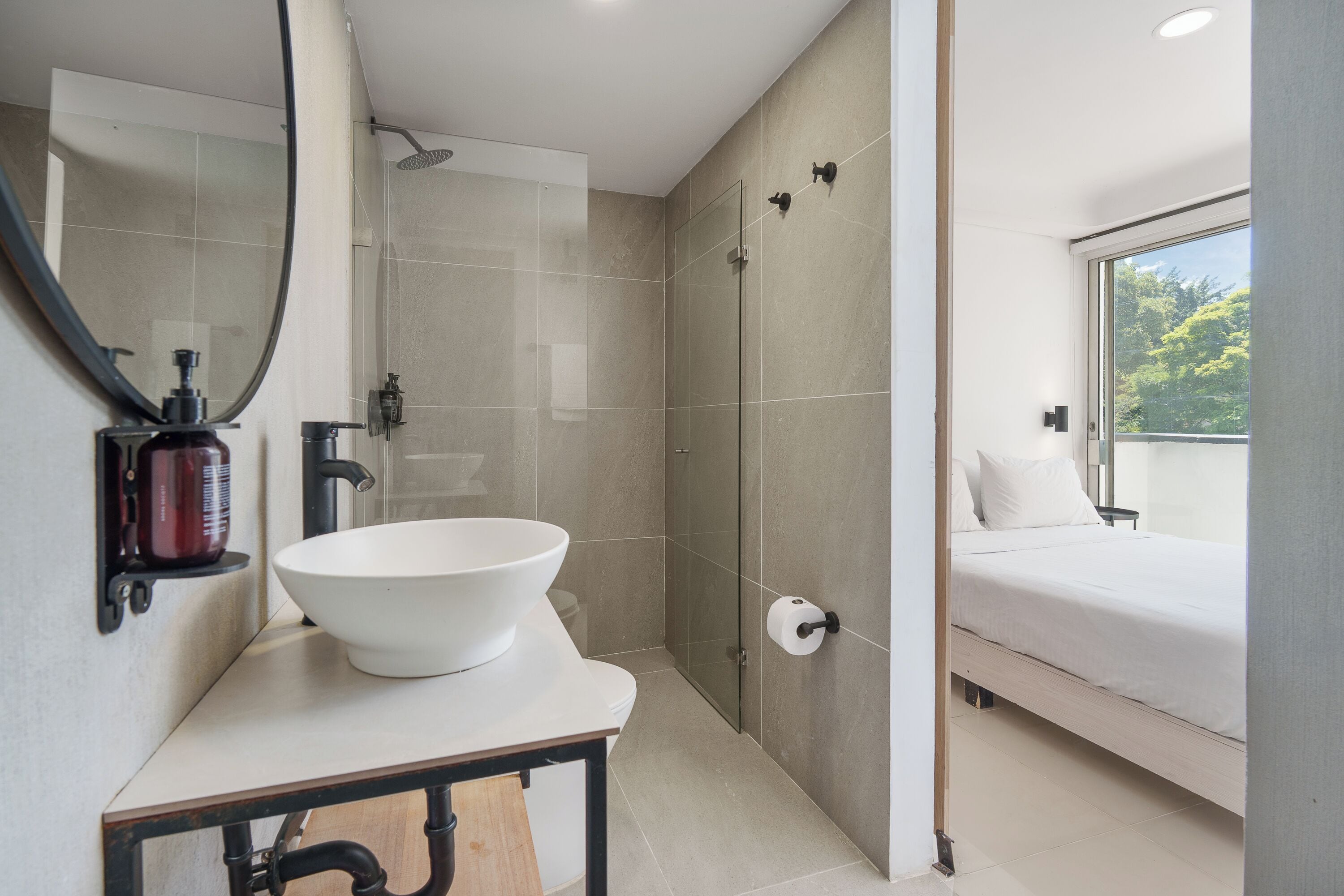 A modern bathroom with a large white sink, a glass shower enclosure, and a bed with white bedding.