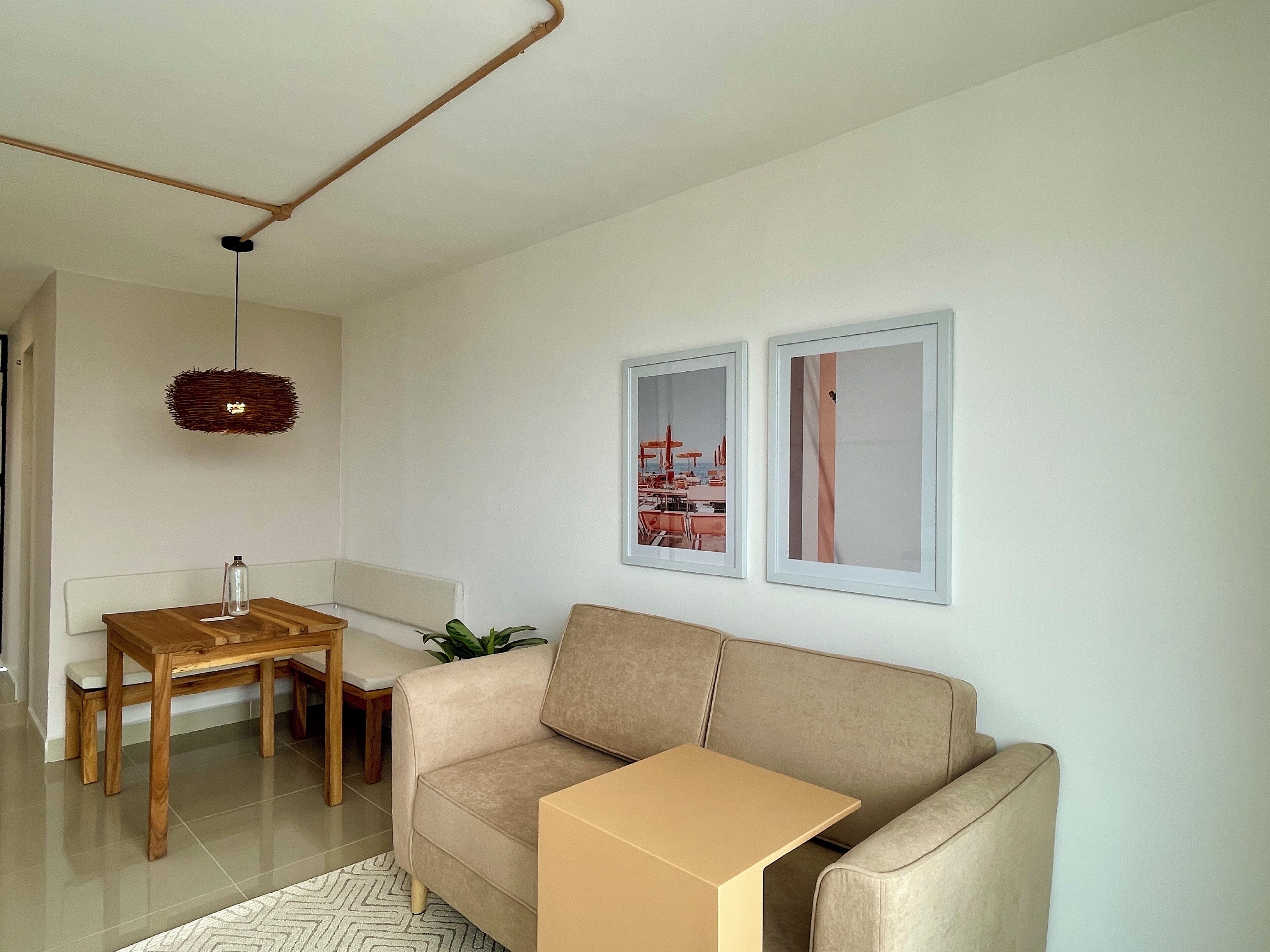 A living room with a beige sofa, a wooden table, and a small plant. There are two framed pictures on the wall.
