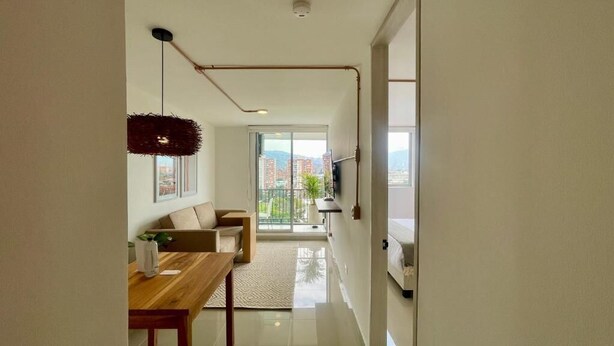 Ein modernes Wohnzimmer mit einer Couch, einem Couchtisch und einem Fernseher. Eine Essecke mit Holztisch und Stühlen ist ebenfalls zu sehen. Das Zimmer hat große Fenster mit Blick auf die Stadt.