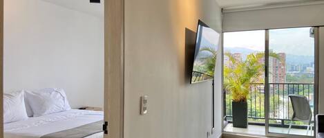 A modern hotel room with a bed, a TV mounted on the wall, a balcony with a view of the city, and a potted plant.