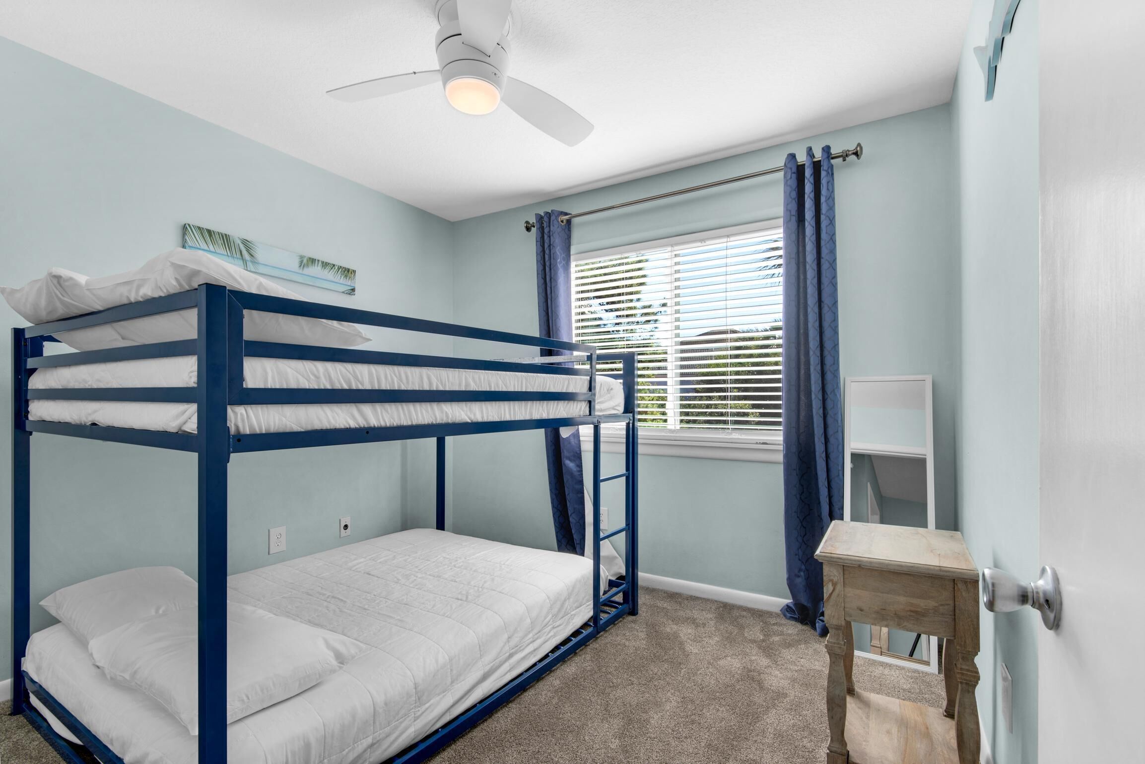 A bunk bed room with a ceiling fan, a window with blinds, and a small wooden table.