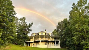 A two-story house with a wrap-around porch, surrounded by lush greenery and a rainbow in the sky. - The Stillwater Suite, named after the serene stillness of its surroundings and the moments where the lake is as still as glass. (Jasper)
