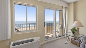 A room with a large window overlooking a beach, a white air conditioner, a glass table with a lamp, and a chair.