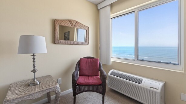 A room with a view of the ocean, a chair, a table with a lamp, and a wall-mounted mirror.