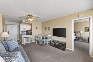 Ein Wohnzimmer mit einer Couch, einem Esstisch mit Stühlen, einem Deckenventilator, einer Küchenzeile und einem an der Wand befestigten Fernseher.