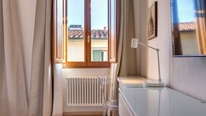 A room with a wooden floor, a white radiator, a glass table, a white desk, a lamp, and a window with curtains. - Mamo Florence - Sacha Apartment (Florence)