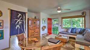 A living room with a glass table, wooden chairs, a sofa with patterned cushions, a red door, and a view of greenery outside. - Summer Deal Kapaa 1BR Cottage Walk to Beach Shops and Dining (KAPAA)