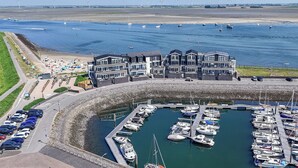 A marina with boats docked, a building with multiple balconies, and a sandy beach area with umbrellas.