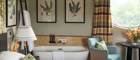 A modern bathroom with a freestanding bathtub, a chair with a towel, framed botanical prints on the wall, and a window with curtains.