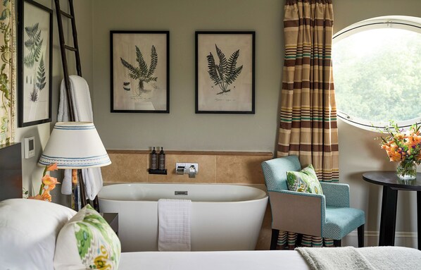 A modern bathroom with a freestanding bathtub, a chair with a towel, framed botanical prints on the wall, and a window with curtains. - Bowood Hotel Spa and Golf Resort (Calne)