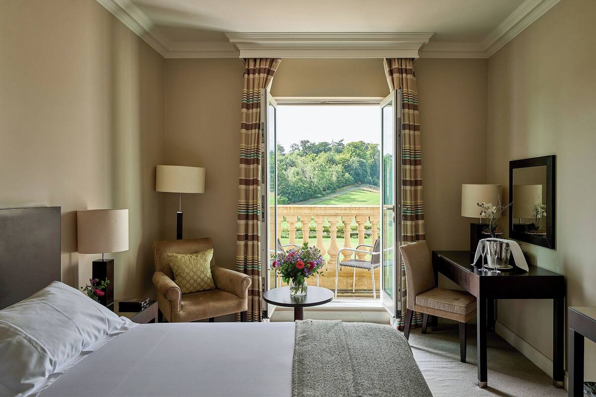 Ein Hotelzimmer mit einem Bett, zwei Sesseln, einem Schreibtisch und Blick auf einen Garten durch ein großes Fenster.