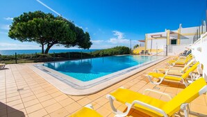 Zona de piscina con vista despejada al mar, un árbol y un edificio de fachada amarilla y blanca.