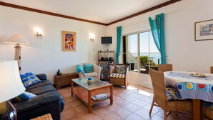 Una sala de estar con un sofá, una mesa de centro y una mesa de comedor con sillas. Hay un televisor sobre un mueble bajo y una estantería.