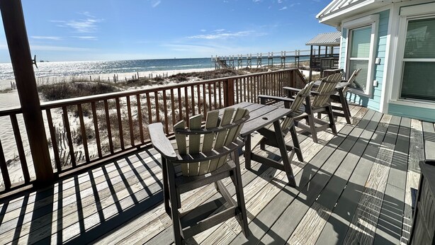 Une terrasse en bois avec des chaises et une table, donnant sur une plage, une clôture en bois et un bâtiment à la façade bleu clair.