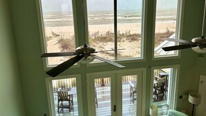 Une chambre dotée de grandes fenêtres donnant sur une plage, d’un ventilateur de plafond et offrant une vue sur une terrasse aménagée avec du mobilier d’extérieur.
