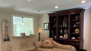 A living room with a floral-patterned sofa, a round coffee table, a wooden cabinet with glassware, and a large window. - Come visit our “Sweet Retreat at the End of the Street” where you can relax!!! (Summerville)
