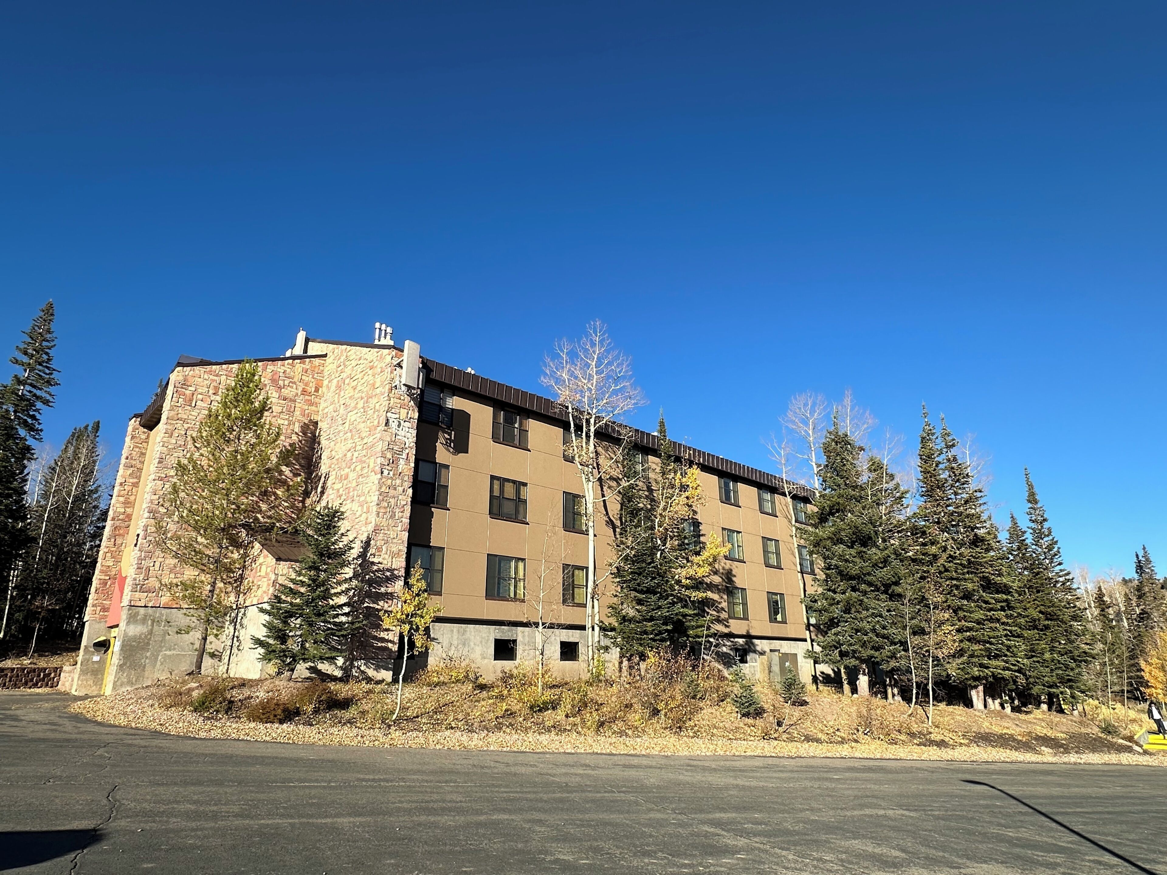 A multi-story building with a stone facade and a flat roof, surrounded by trees and a parking lot.