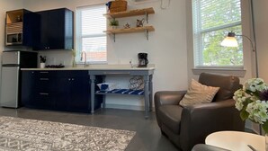 A modern kitchen with dark cabinets, a built-in microwave, and a coffee maker on the counter. There is a dining area with a table and chairs, and a living room with a sofa and a lamp. - Elizabeth Manor in Downtown Huntsville (Huntsville)