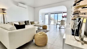 A modern living room with a white sofa, a round woven ottoman, and a flat-screen TV on a white stand.