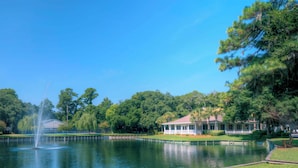 Um lago tranquilo com uma fonte, cercado por vegetação exuberante e uma casa com varanda.